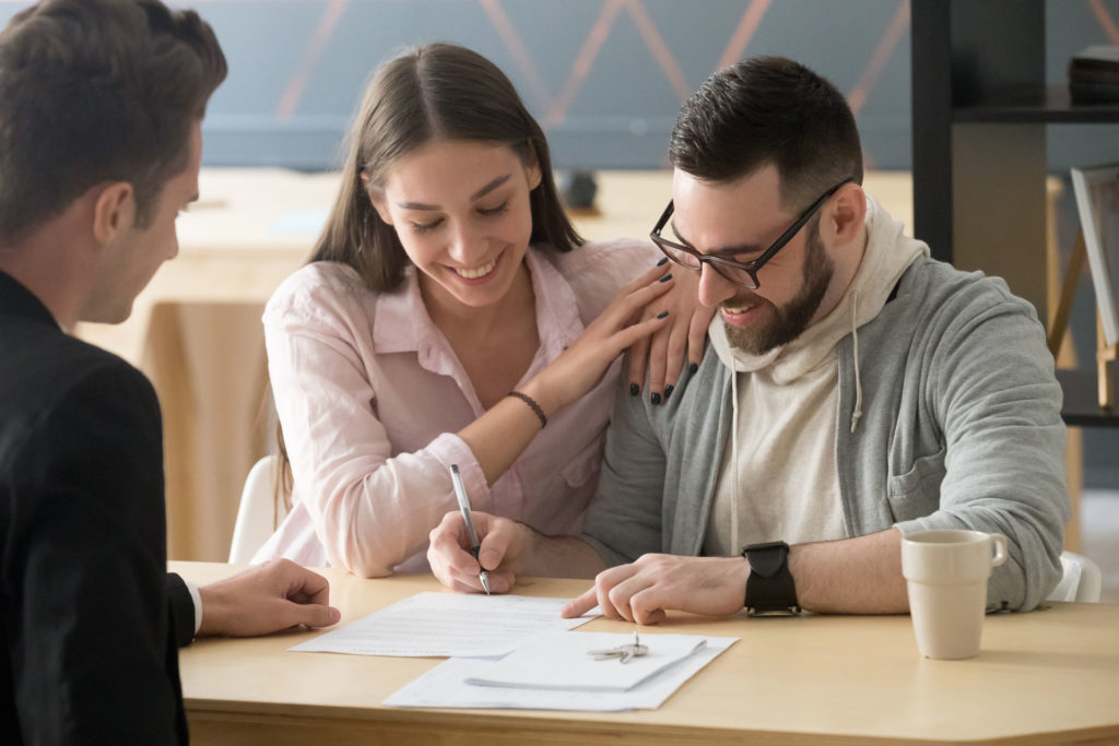 A young couple sitting at a desk and signing documents, with a lawyer overseeing the transaction.